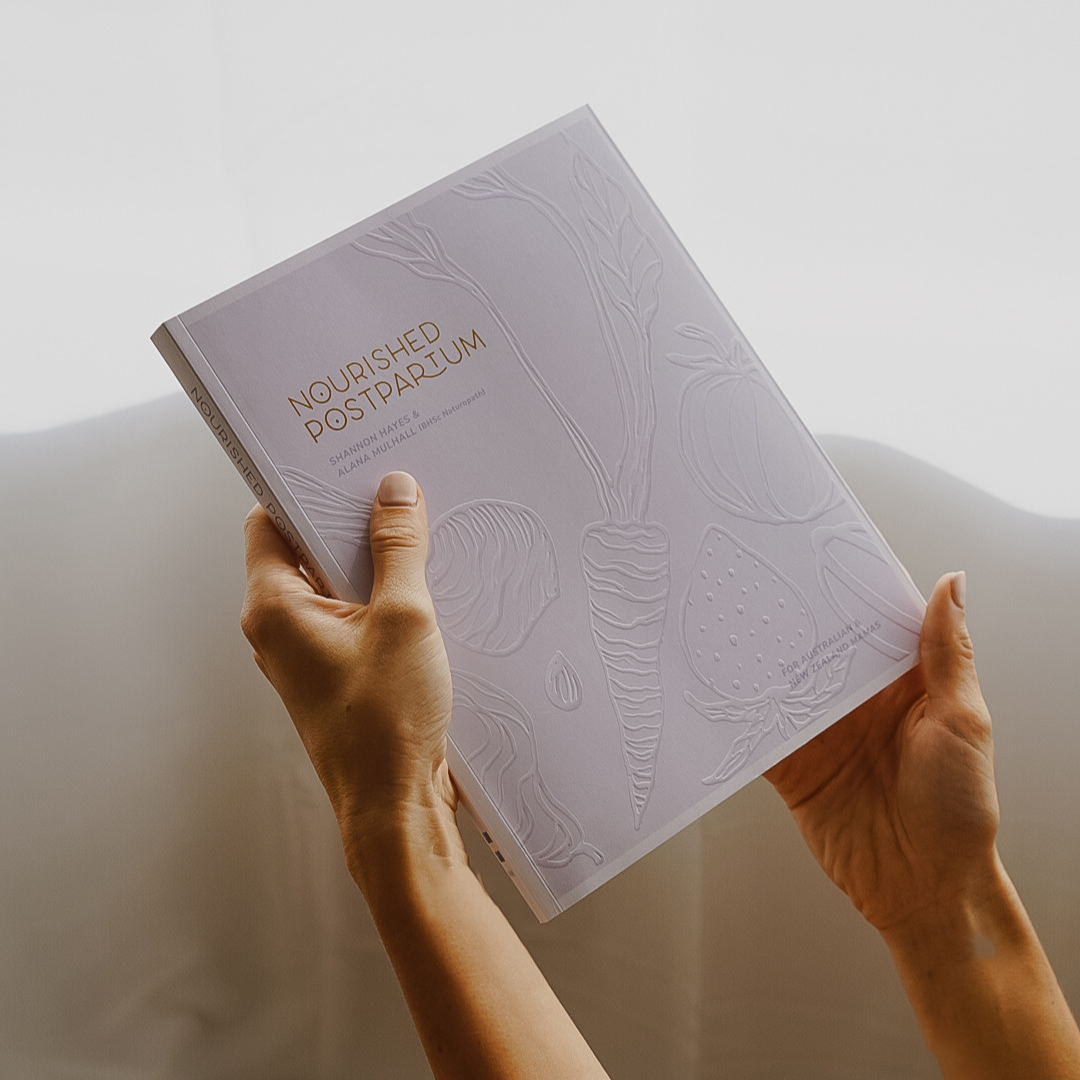 A woman holding up the book 'Nourished Postpartum' by Mothers Mylk.