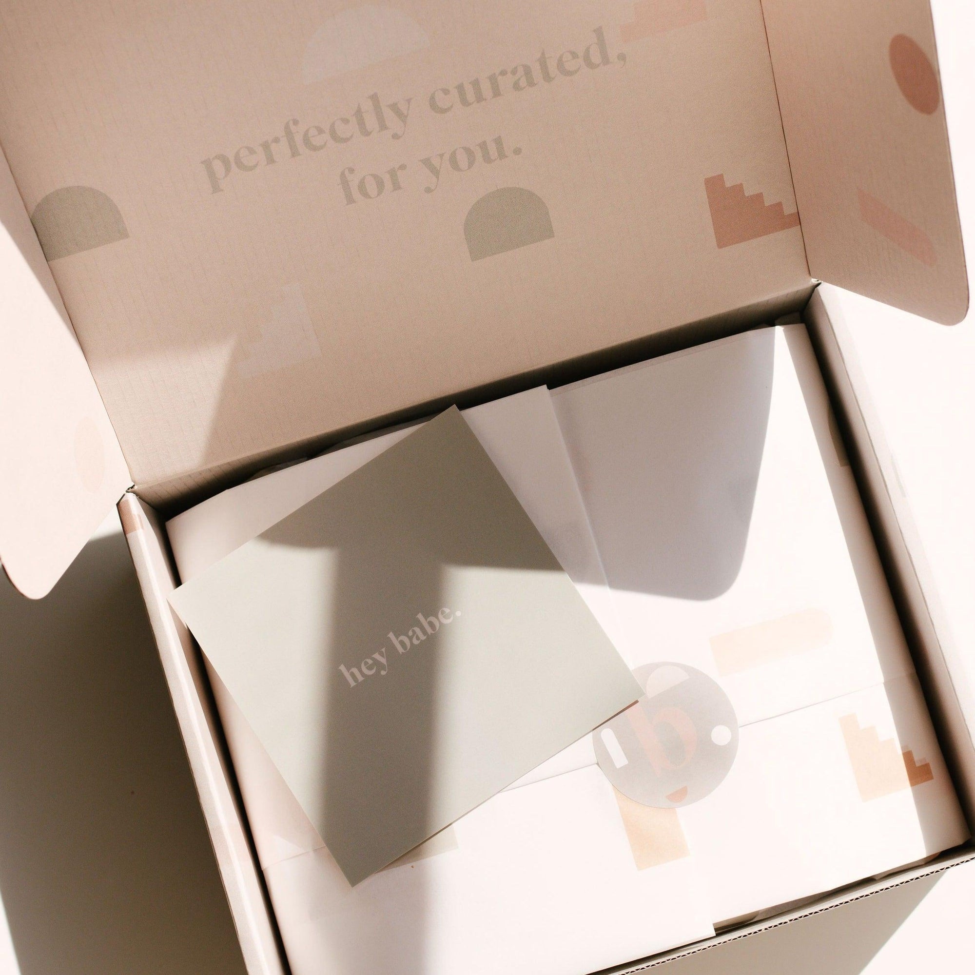 An open cardboard box reveals tissue paper with a small card on top that reads "hey babe | gift card" by biglittlethings. The box lid has the text "perfectly curated for you" printed on the inside.