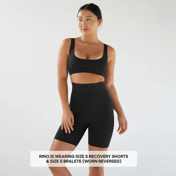 A woman stands in a studio wearing Bare Mum Postpartum Recovery Shorts and a sports bra, with a text overlay stating her clothing sizes and style.