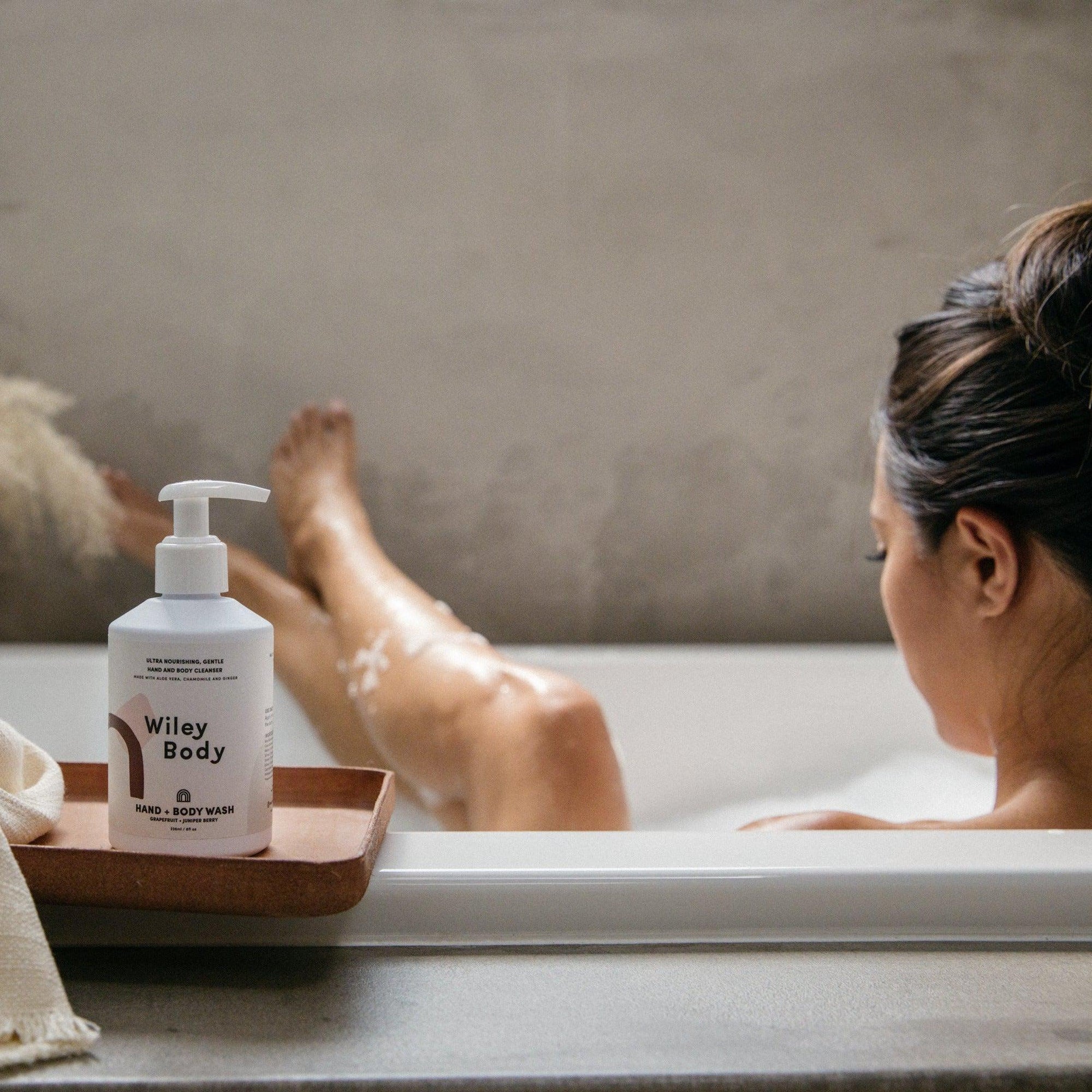 A woman is sitting in a tub with a bottle of Wiley Body hand & body wash.