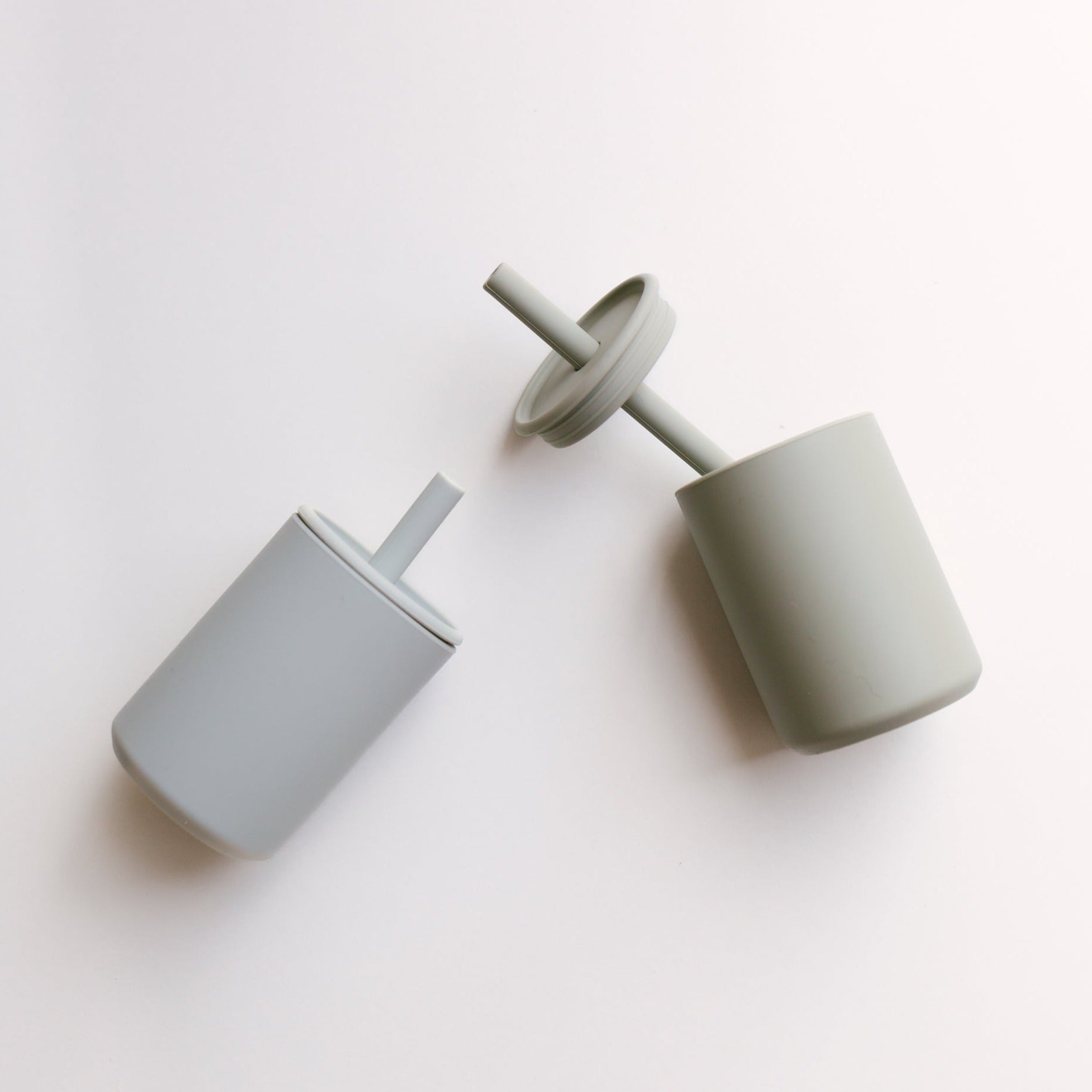 Two gray sippy cups from Rommer, each accompanied by a lid and straw, are positioned diagonally against a plain white background, highlighting their premium silicone dinnerware quality.