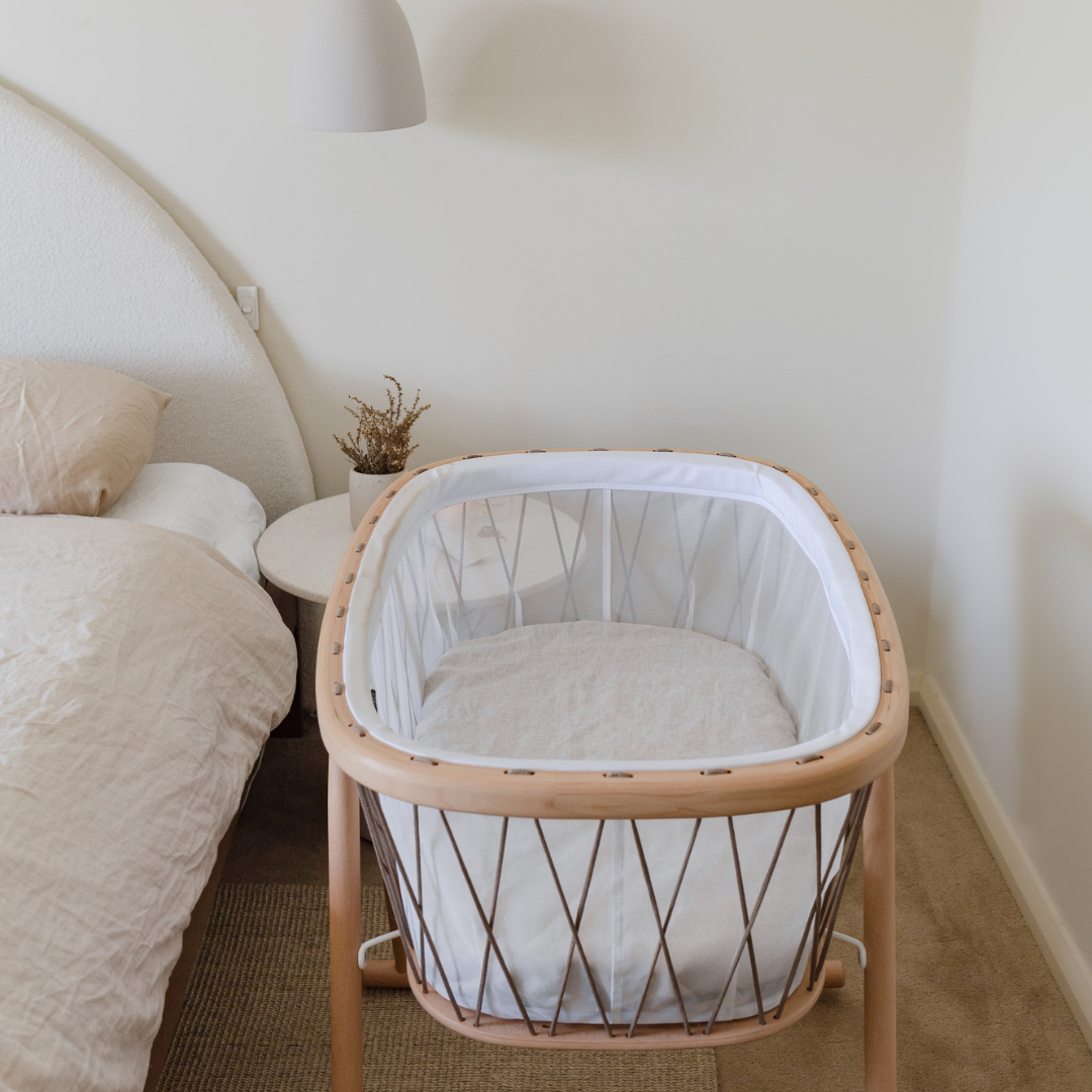 A Warren Hill baby cot with a French linen fitted bassinet sheet in natural in a bedroom with a bed.